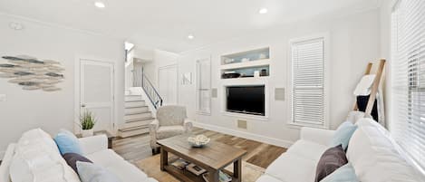 A modern living room with a white sofa, a coffee table, and a TV. There is a staircase in the background leading to another level.
