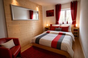 Une chambre avec un grand lit, un fauteuil rouge, un mur en bois, un miroir et une fenêtre avec des rideaux rouges.