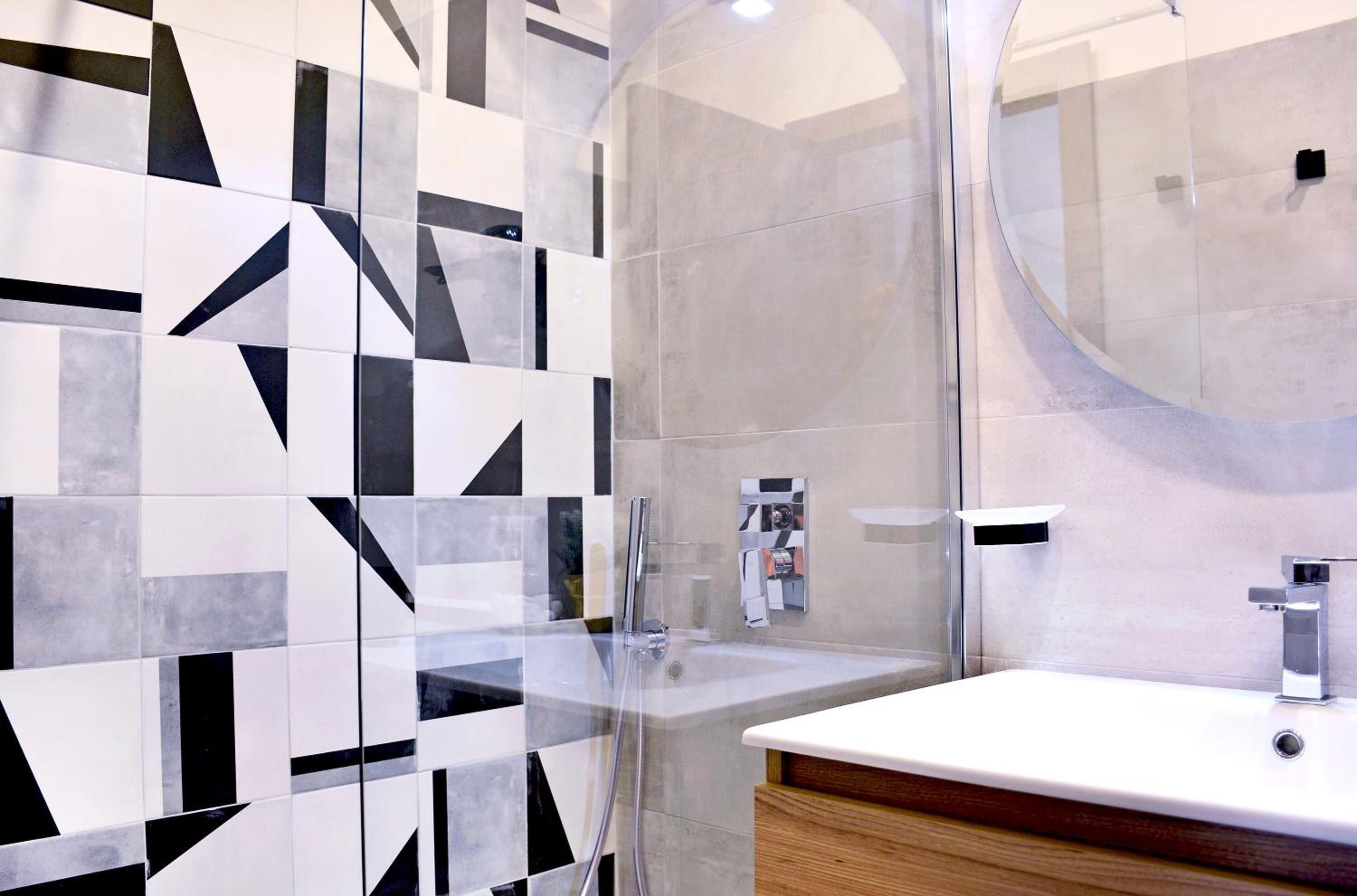 A modern bathroom with a geometric tile backsplash, a glass shower enclosure, and a wooden vanity with a white sink and chrome fixtures.