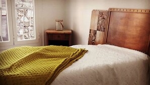 Un letto con piumone bianco e coperta gialla, un comodino in legno con un vasetto e una testiera decorata.