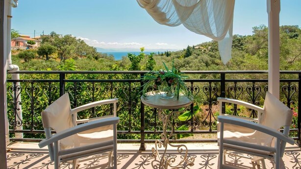 Ein Balkon mit zwei weißen Stühlen, einem kleinen runden Tisch und Blick auf eine Küstenlandschaft.