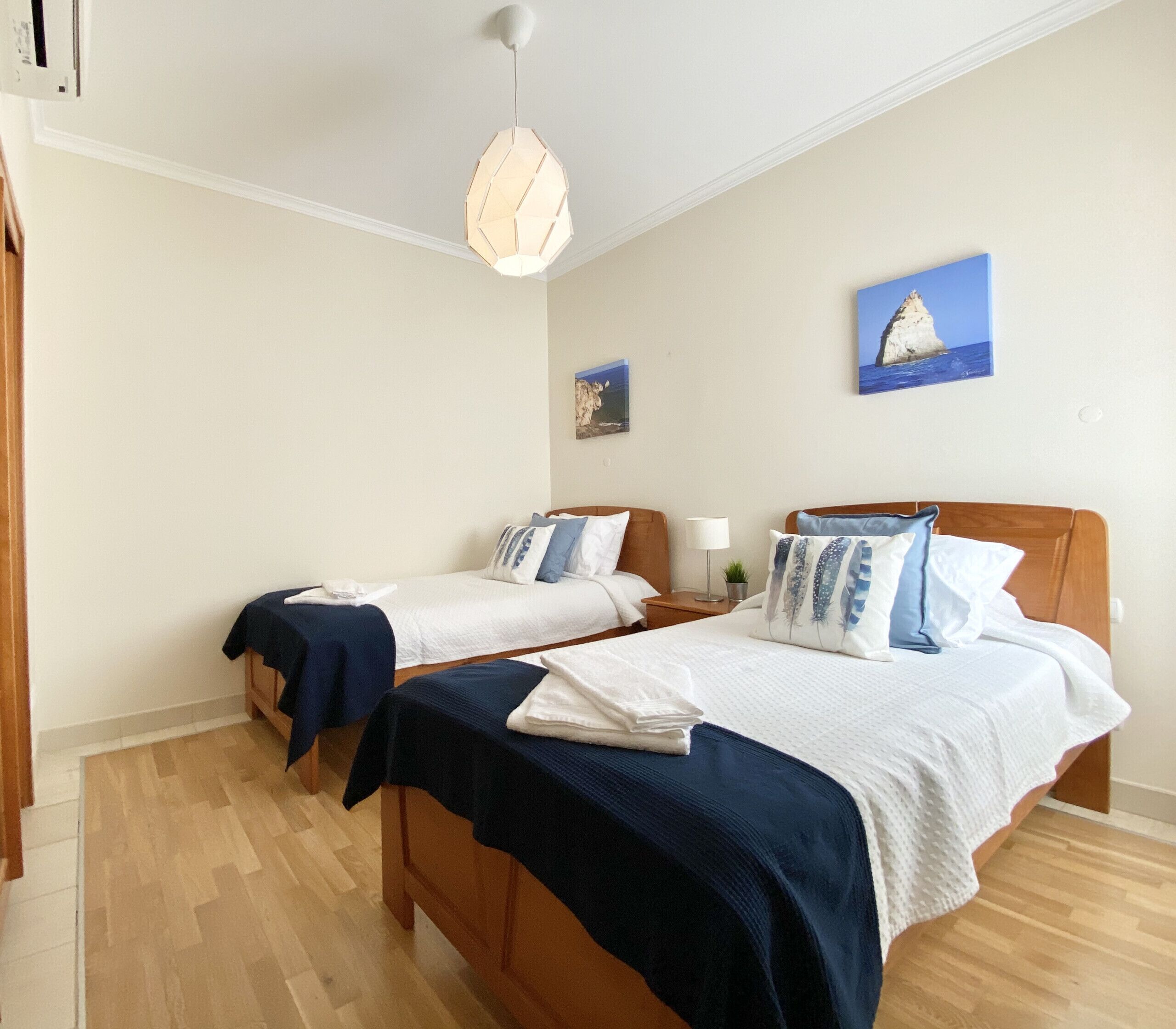 Ein Hotelzimmer mit zwei Betten, Holz-Kopfteilen, einer Pendelleuchte und einem Gemälde einer Küstenlandschaft.