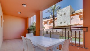 Een balkon met een witte tafel en stoelen, uitzicht op een gebouw met balkons, en een palmboom.