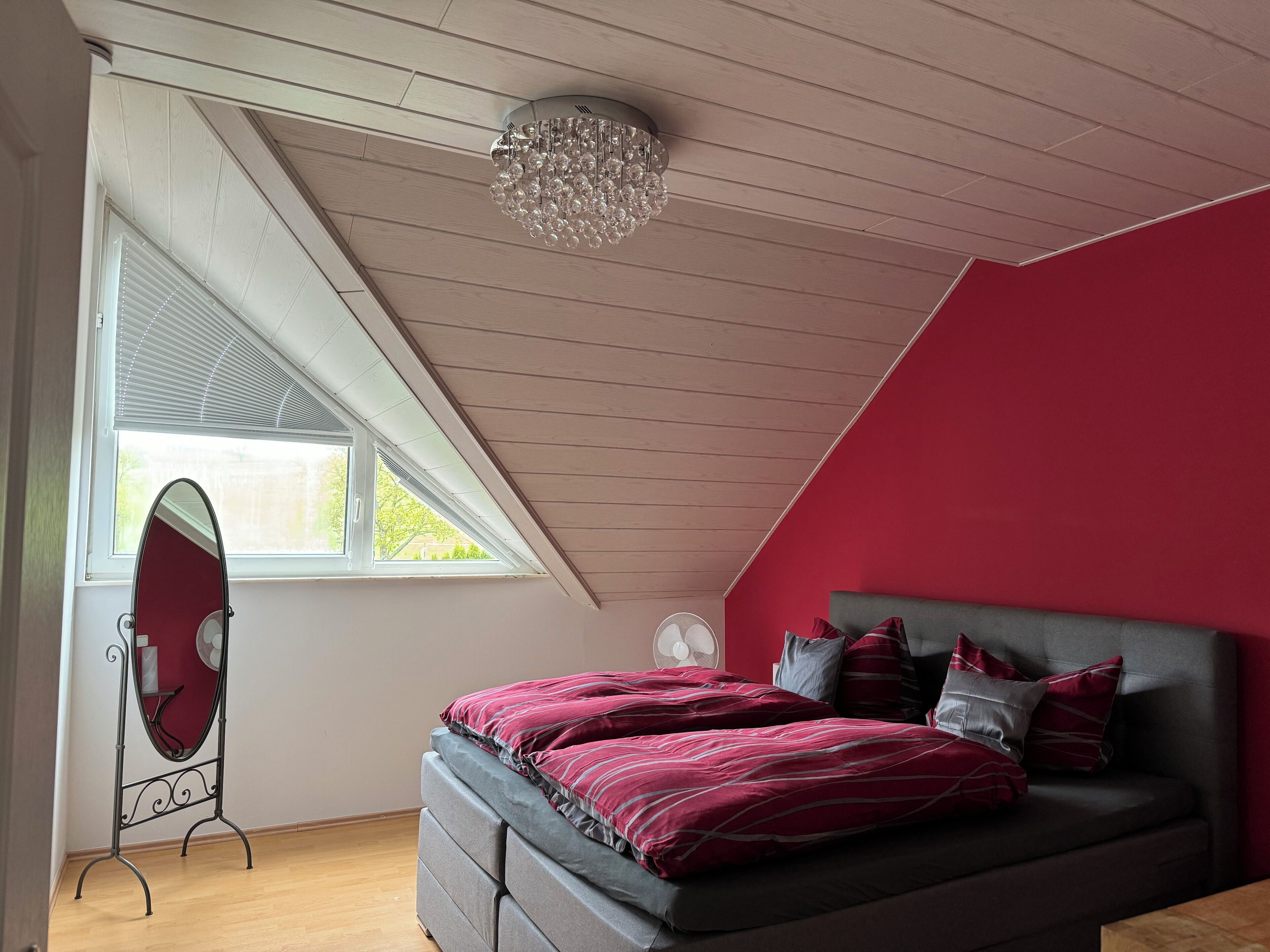 Une chambre avec un plafond en pente, un lit avec une literie rouge et grise, un grand miroir et une fenêtre avec des stores.
