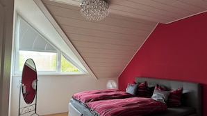 Ein Schlafzimmer mit schrägen Wänden, einem Bett mit roter und grauer Bettwäsche, einem stehenden Spiegel und einem Fenster mit Jalousien.