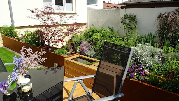 Un giardino con una terrazza in legno, una sedia e un tavolo con un vaso di fiori.