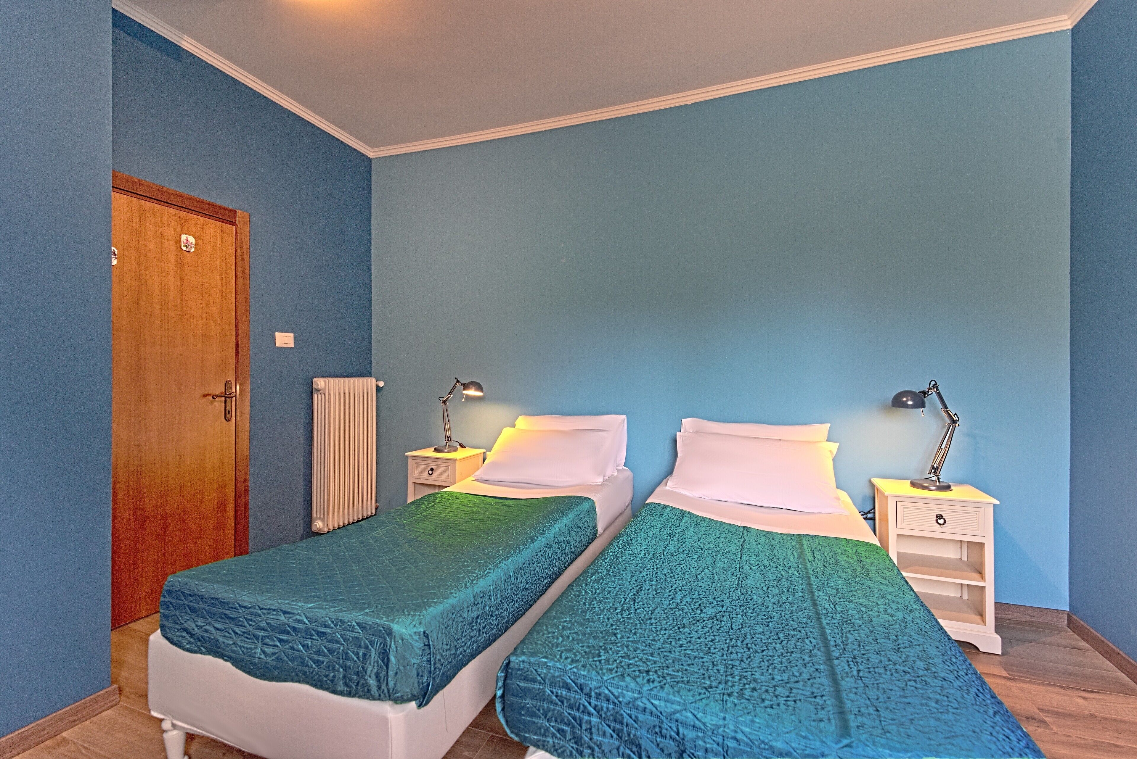 A room with two single beds, a wooden door, a white radiator, and a nightstand with a lamp.