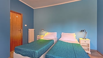A room with two single beds, a wooden door, a white radiator, and a nightstand with a lamp.