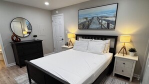 A bedroom with a large bed, two bedside tables with lamps, a dresser, a mirror, and a painting on the wall. - Ocean Block Gem in North Ocean City (Ocean City)