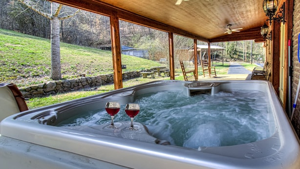 A hot tub with two glasses of red wine, surrounded by stone walls and wooden beams, with a view of a grassy yard and a pathway leading to a house. - Kenny's Mountain Hideaway w/ Hot Tub, Arcade, Fireplace, Views, & 10 King Beds (Sevierville Tennessee)