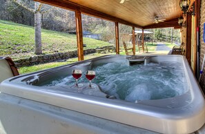 A hot tub with two glasses of red wine, surrounded by stone walls and wooden beams, with a view of a grassy yard and a pathway leading to a house. - Kenny's Mountain Hideaway w/ Hot Tub, Arcade, Fireplace, Views, & 10 King Beds (Sevierville Tennessee)