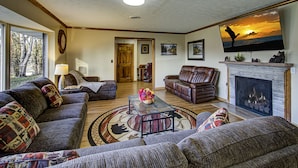 A living room with a fireplace, leather sofas, a coffee table with a floral centerpiece, and a large TV mounted above the fireplace. - Kenny's Mountain Hideaway w/ Hot Tub, Arcade, Fireplace, Views, & 10 King Beds (Sevierville Tennessee)