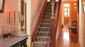Un escalier orné de balustrades travaillées et surmonté d’un lustre, menant à une pièce dotée d’un plancher en bois et d’un tapis à motifs.