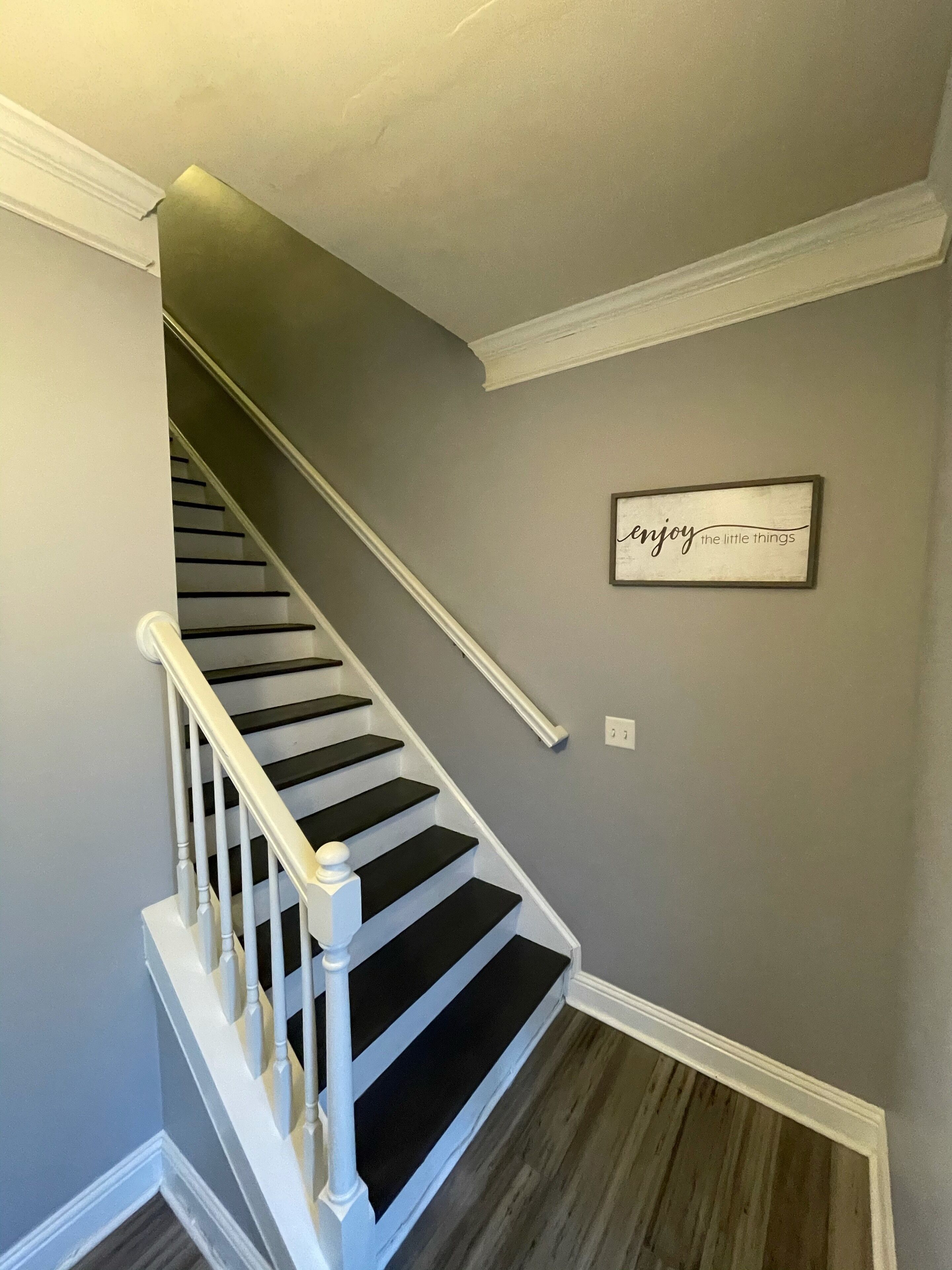 Un escalier avec des rampes blanches et des marches noires, menant à un palier où est fixée une plaque murale portant l’inscription : « appréciez les petites choses de la vie ».