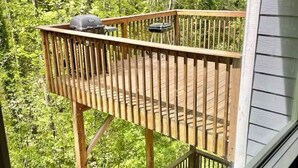 Una terraza de madera con una parrilla y una silla, rodeada de abundante follaje verde.