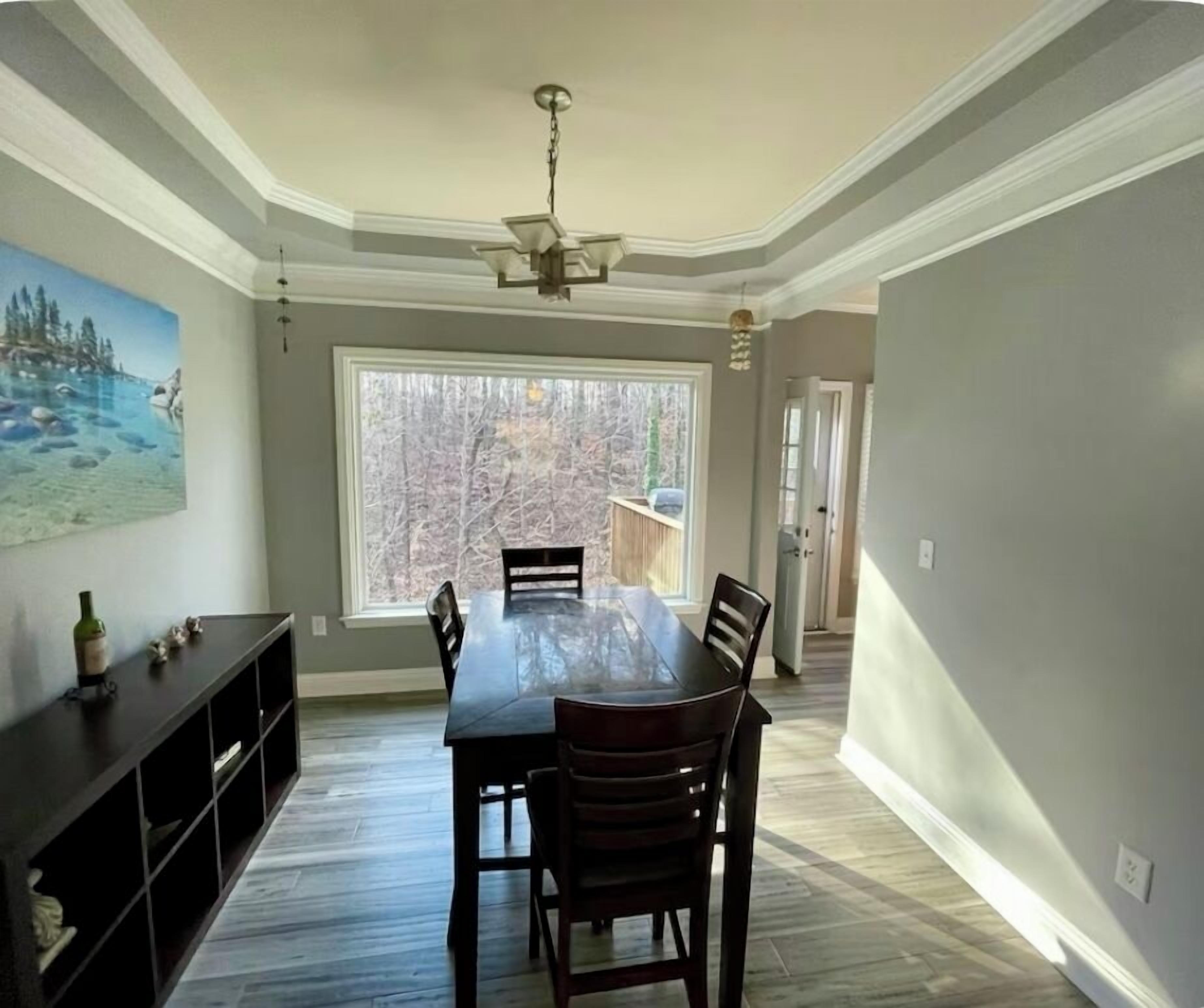 Une salle à manger avec une grande table et des chaises, un ventilateur de plafond, un tableau au mur et une vue sur l’extérieur.