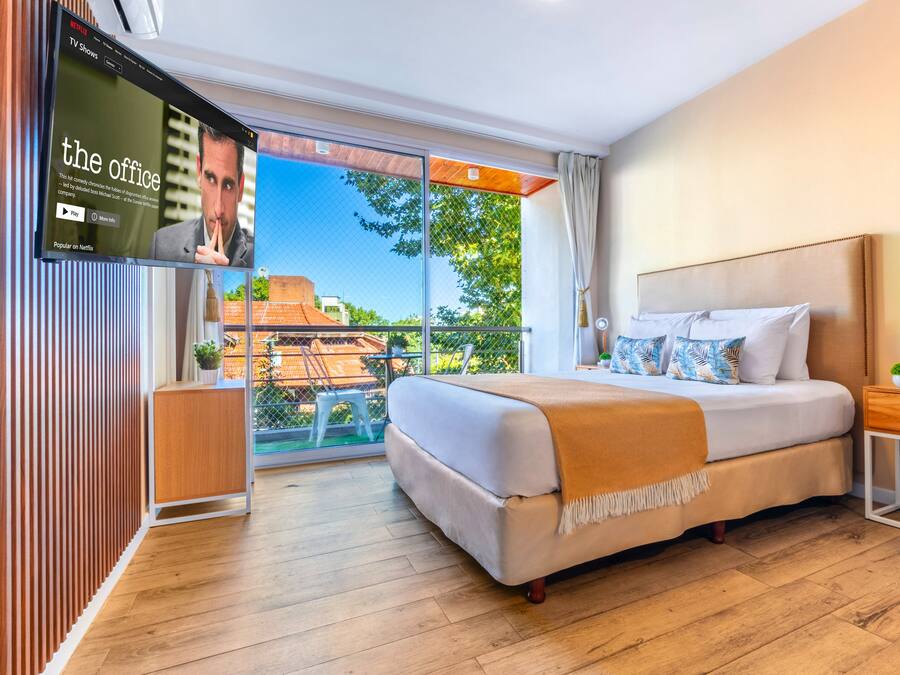 A modern hotel room with a large bed, a TV mounted on the wall, a balcony with a view, and a wooden floor.