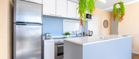 A modern kitchen with a stainless steel refrigerator, white cabinets, a central island, hanging plants, and a microwave above the island.