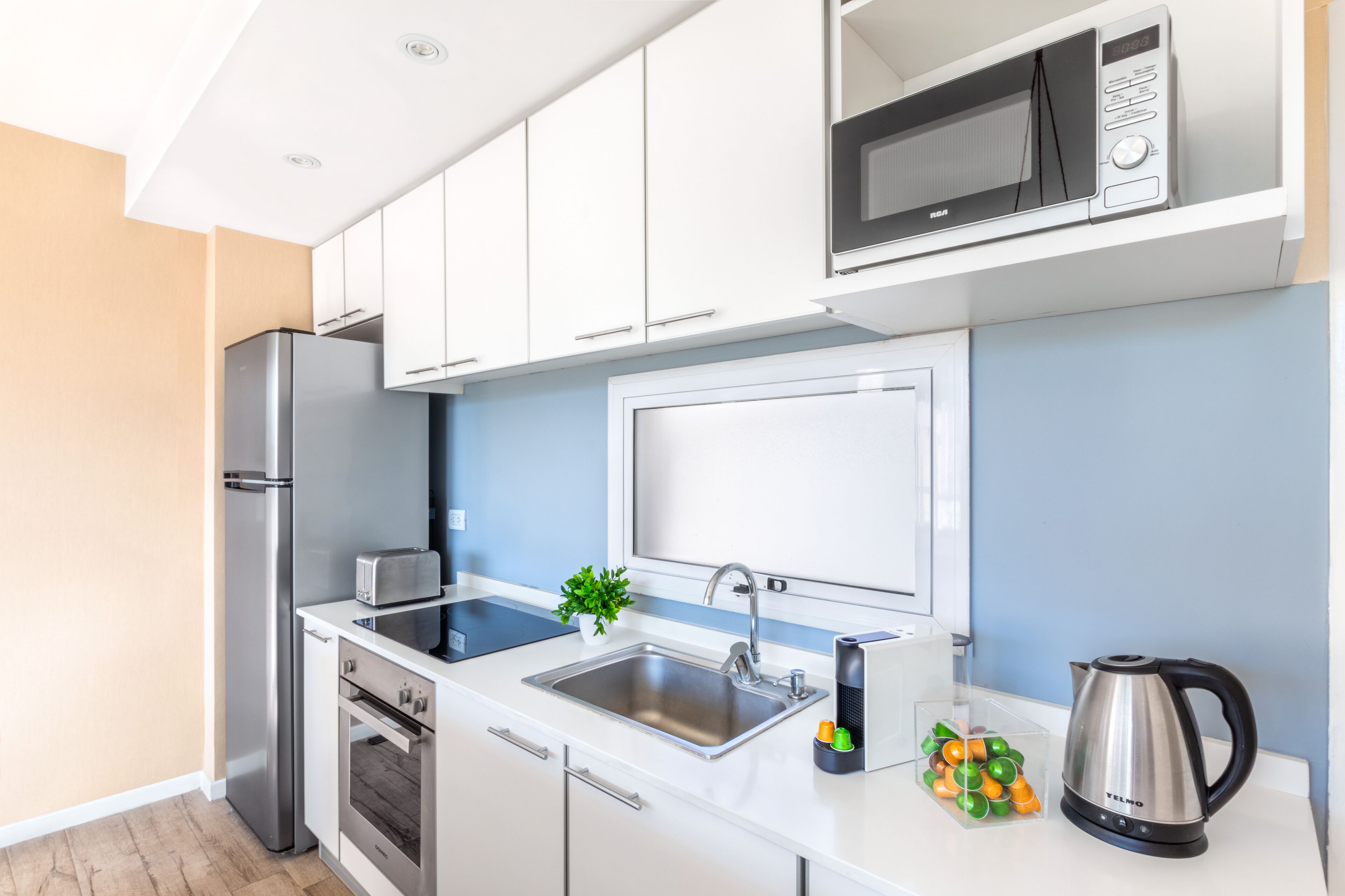 Una cocina moderna con armarios blancos, un refrigerador de acero inoxidable, un microondas, un fregadero y una tetera.