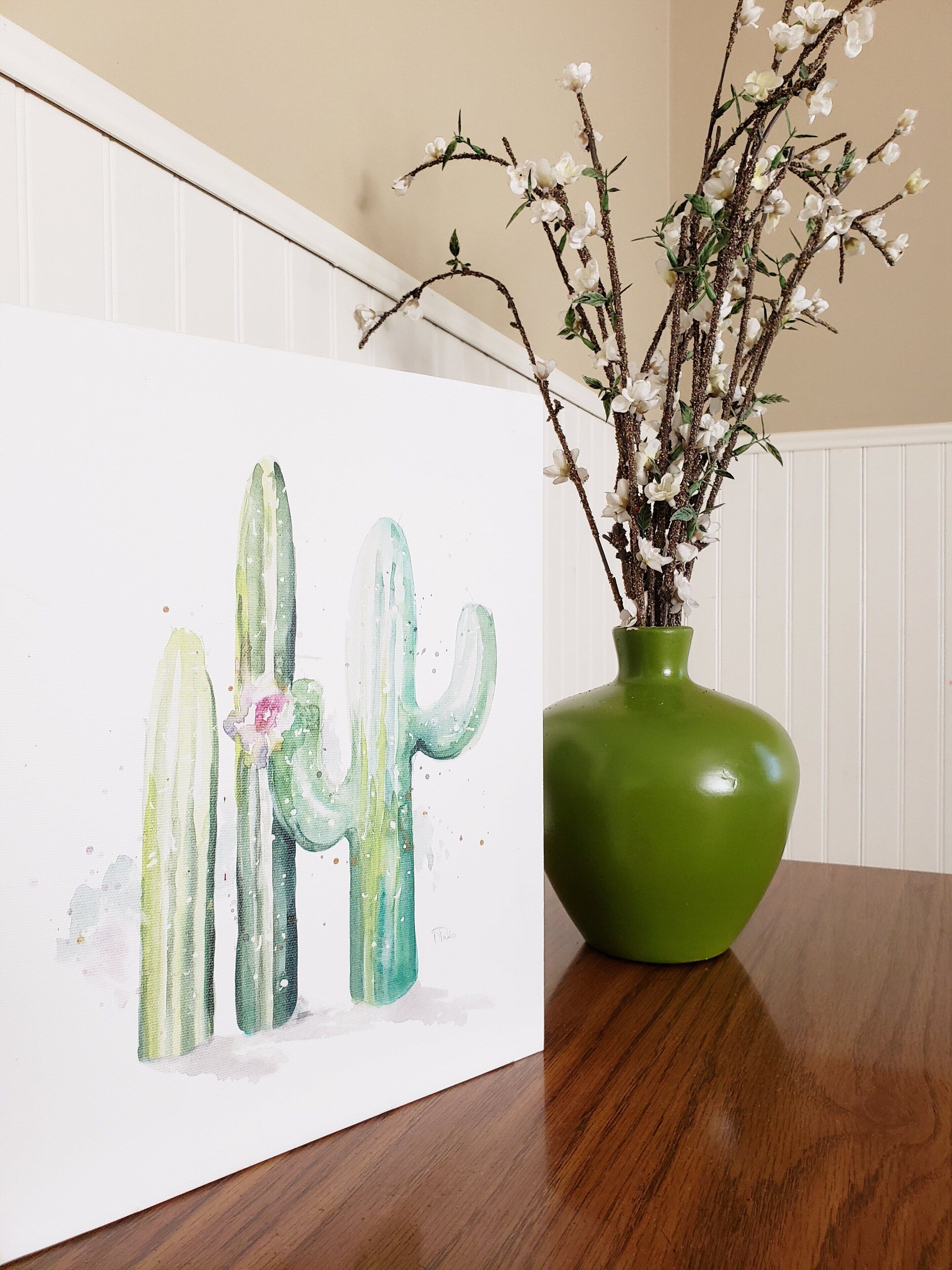 A cactus painting on a wall next to a green vase with branches and flowers.