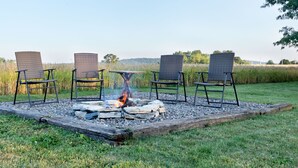 A fire pit with four chairs around it, set in a grassy area with a field in the background.