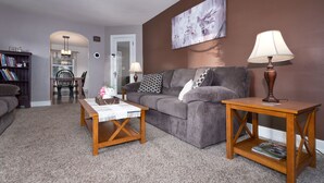 A living room with a grey sofa, wooden coffee table, and side tables, a bookshelf, and a painting on the wall.