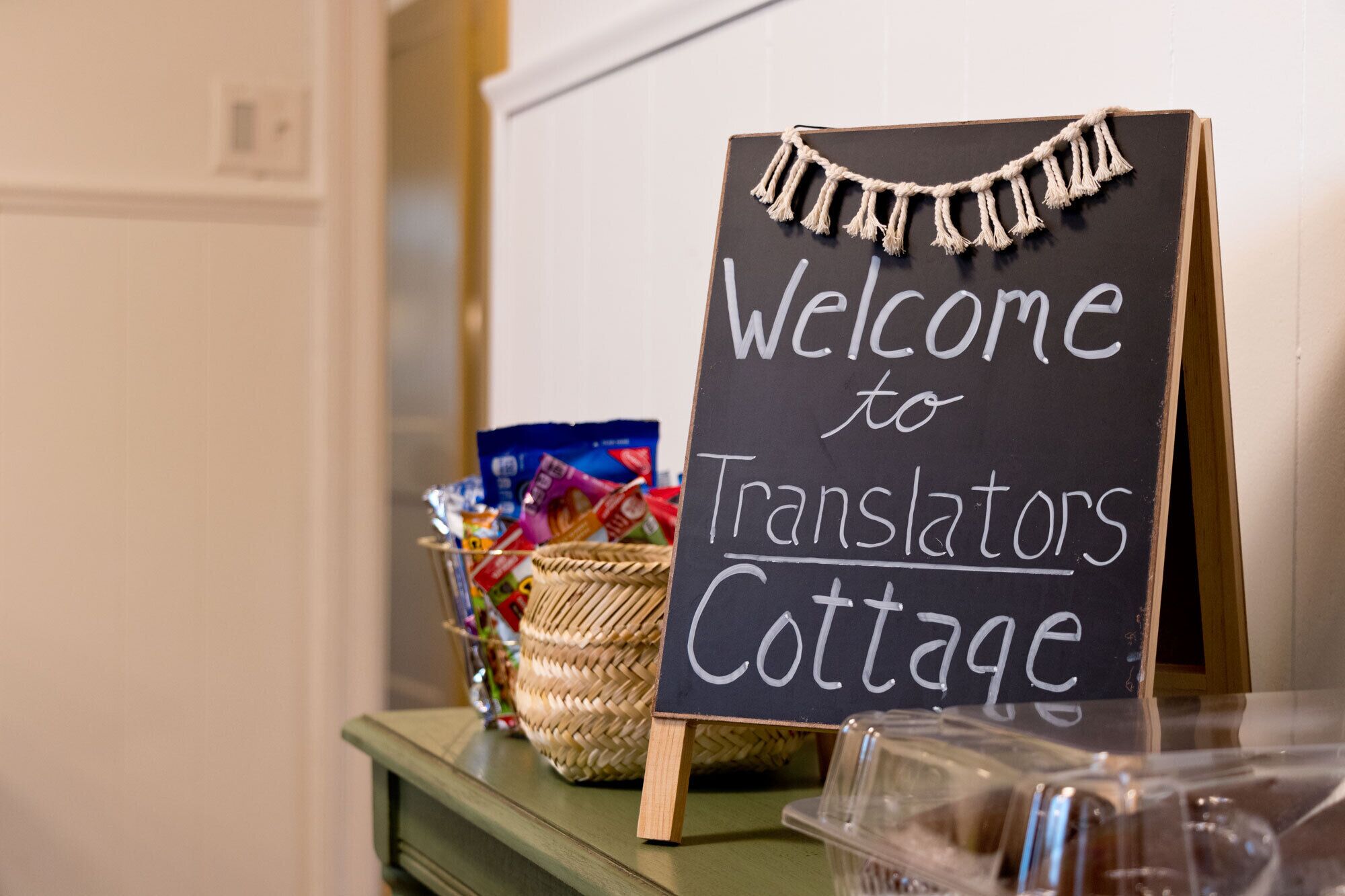 Un cartello con la scritta "Welcome to Translators Cottage", un cesto di vimini e un contenitore di plastica trasparente su un tavolo di legno.