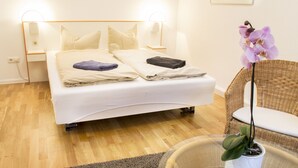 Un lit bien fait, avec des oreillers et des couvertures, une chaise en osier, une table en verre ornée d’une orchidée en pot et une lampe fixée au mur.