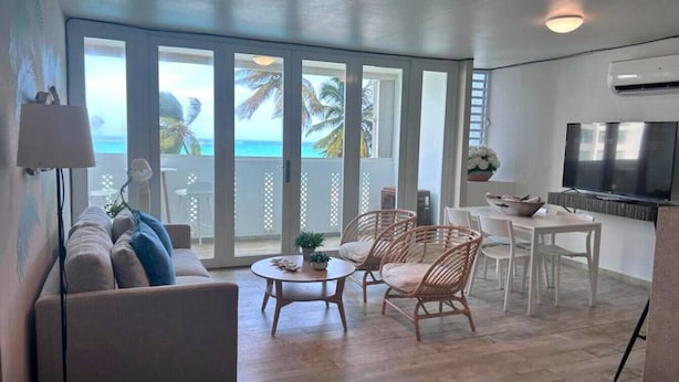 A spacious living area with a sofa, round coffee table, wicker chairs, and a dining table. A large TV is mounted on the wall, and there is a view of the ocean through large sliding doors. - Brisa Oceanfront + Beach + Pool + Ocean View Balcony (San Juan)