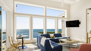 Ein Wohnzimmer mit großem Fenster zum Meer, einer Couch, einem Couchtisch und einem an der Wand befestigten Fernseher.