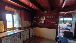 A kitchen with a wooden ceiling, a sink, and a window with curtains. There are shelves with glasses and a framed picture. - Penthouse Casa Orymar El Cotillo (El Cotillo)