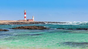 A lighthouse with a red and white striped top, situated on a rocky shoreline with clear turquoise water. - Penthouse Casa Orymar El Cotillo (El Cotillo)