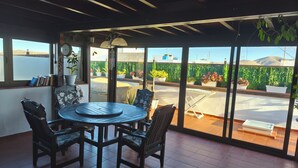 A sunlit patio with a round table and chairs, large windows with a view of a green hedge and mountains, and a ceiling with wooden beams. - Penthouse Casa Orymar El Cotillo (El Cotillo)