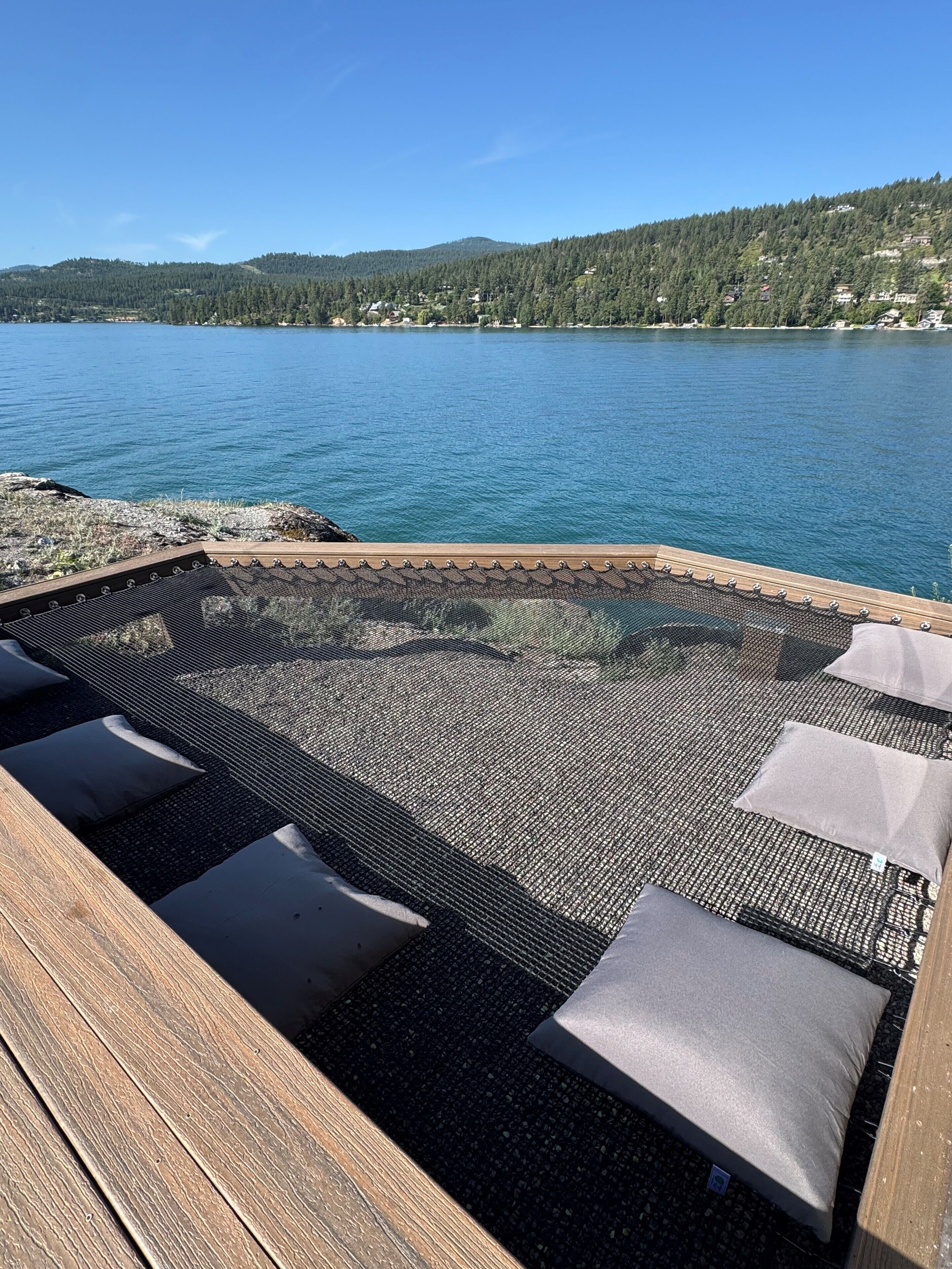 A wooden deck with a woven floor mat and cushions, overlooking a lake and forested hills.