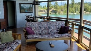 A spacious room with a wooden floor, a sofa with patterned cushions, a round table, and large windows offering a view of a lake and mountains. - PRIVATE ISLAND RETREAT On Gorgeous Flathead Lake Montana!!! (Somers)