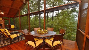 Eine Holzterrasse mit rundem Tisch und Stühlen, umgeben von großen Fenstern mit Blick auf einen Wald.