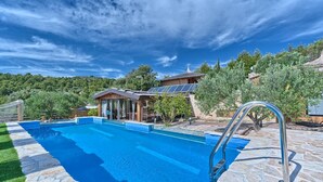 A swimming pool with a stone deck, a wooden house with a glass door, and a view of a forested hillside. - Beachfront Villa New Horizons II (Murter)