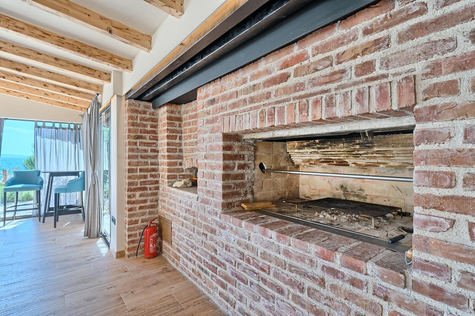 Une cheminée en briques surmontée d’une hotte métallique, un extincteur et une table avec des chaises, le tout près d’une grande fenêtre.
