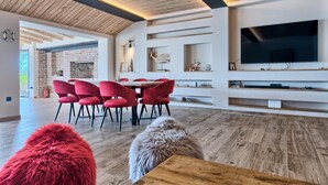 A modern living room with a brick fireplace, red chairs, a wooden dining table, and built-in shelves with decorative items. - Beachfront Villa New Horizons II (Murter)
