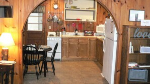 A cozy kitchen with wooden walls, a stove, a sink, and a dining area with a table and chairs. - Three Cub Cottage in historic Flat Rock (Flat Rock)