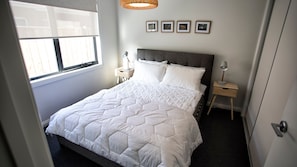 A neatly made bed with a white quilt, a bedside table with a lamp, and a window with blinds.