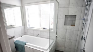 A modern bathroom with a bathtub, a glass shower enclosure, and a window with blinds. - Nautilus in The Point (Point Lonsdale)