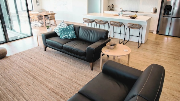 A modern living room with a black leather sofa, a round coffee table, and a kitchen area with a stainless steel refrigerator and bar stools.
