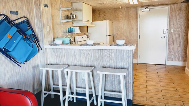 A compact kitchen with a white countertop, a refrigerator, and a sink. There are three white stools and a red chair. A blue suitcase is visible on the left. - Across the street from the beach and a block from Mainstreet (North Myrtle Beach)