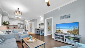 A modern living room with a large TV displaying a beach scene, a cozy seating area, and a kitchen in the background.