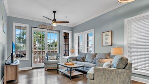 A spacious living room with a ceiling fan, a sofa, a chair, and a coffee table. There are large windows with blinds and a door labeled "BEACH HOUSE".