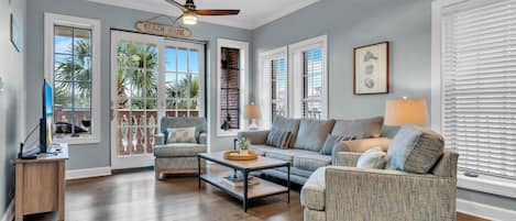 Un salon spacieux doté d’un ventilateur de plafond, d’un canapé, d’un fauteuil et d’une table basse. De grandes fenêtres, équipées de stores, et une porte portant l’inscription « MAISON DE PLAGE » se trouvent dans la pièce.