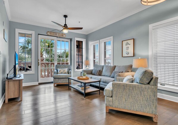 A spacious living room with a ceiling fan, a sofa, a chair, and a coffee table. There are large windows with blinds and a door labeled "BEACH HOUSE". - Stunning Village Condo, Overlooking 30A, Beautifully Decorated Recently Updated (Seacrest Beach)