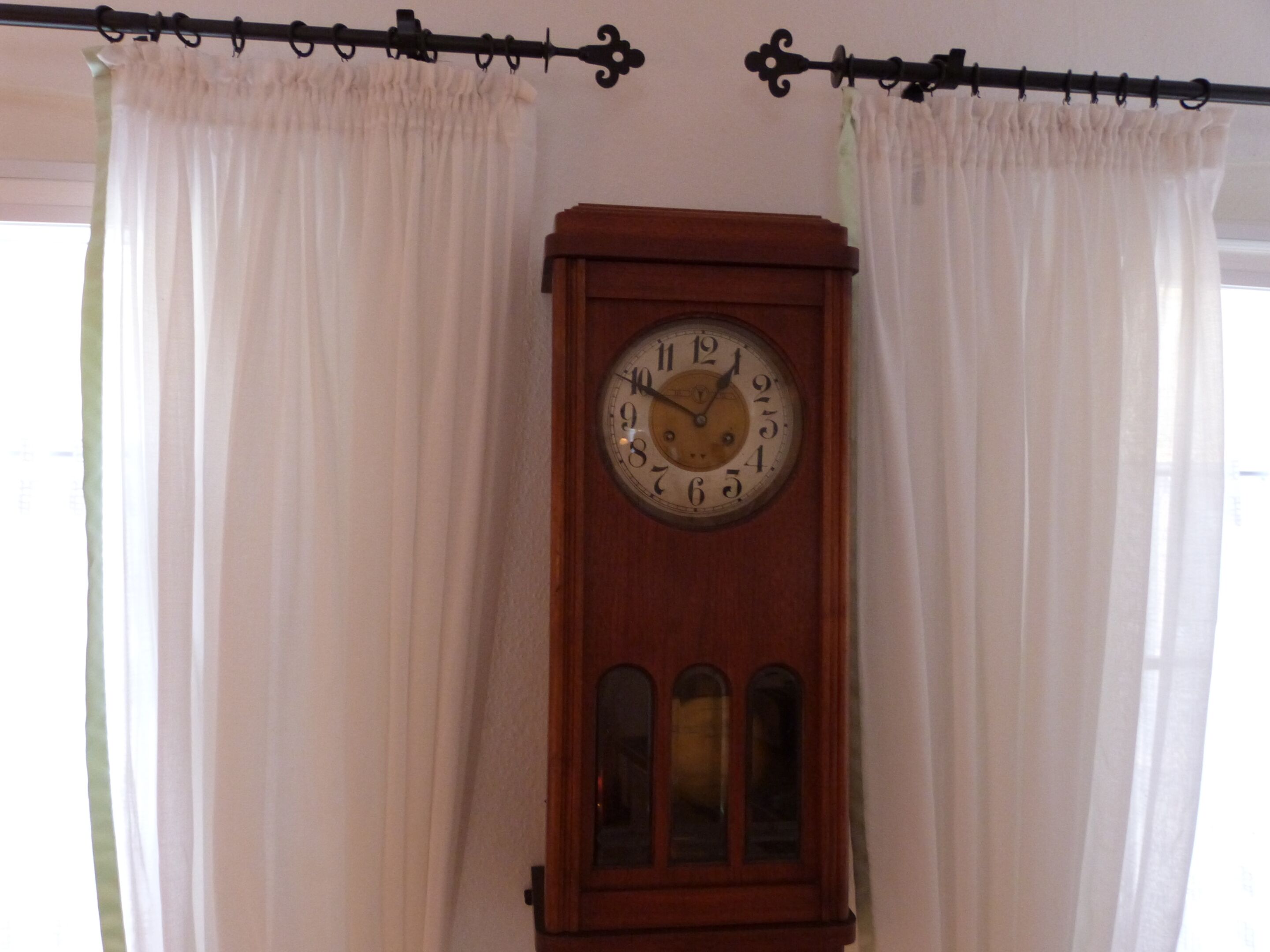 Un reloj de pie de madera con esfera redonda y tres paneles de vidrio debajo, sobre un fondo de cortinas blancas y una barra de cortina negra.