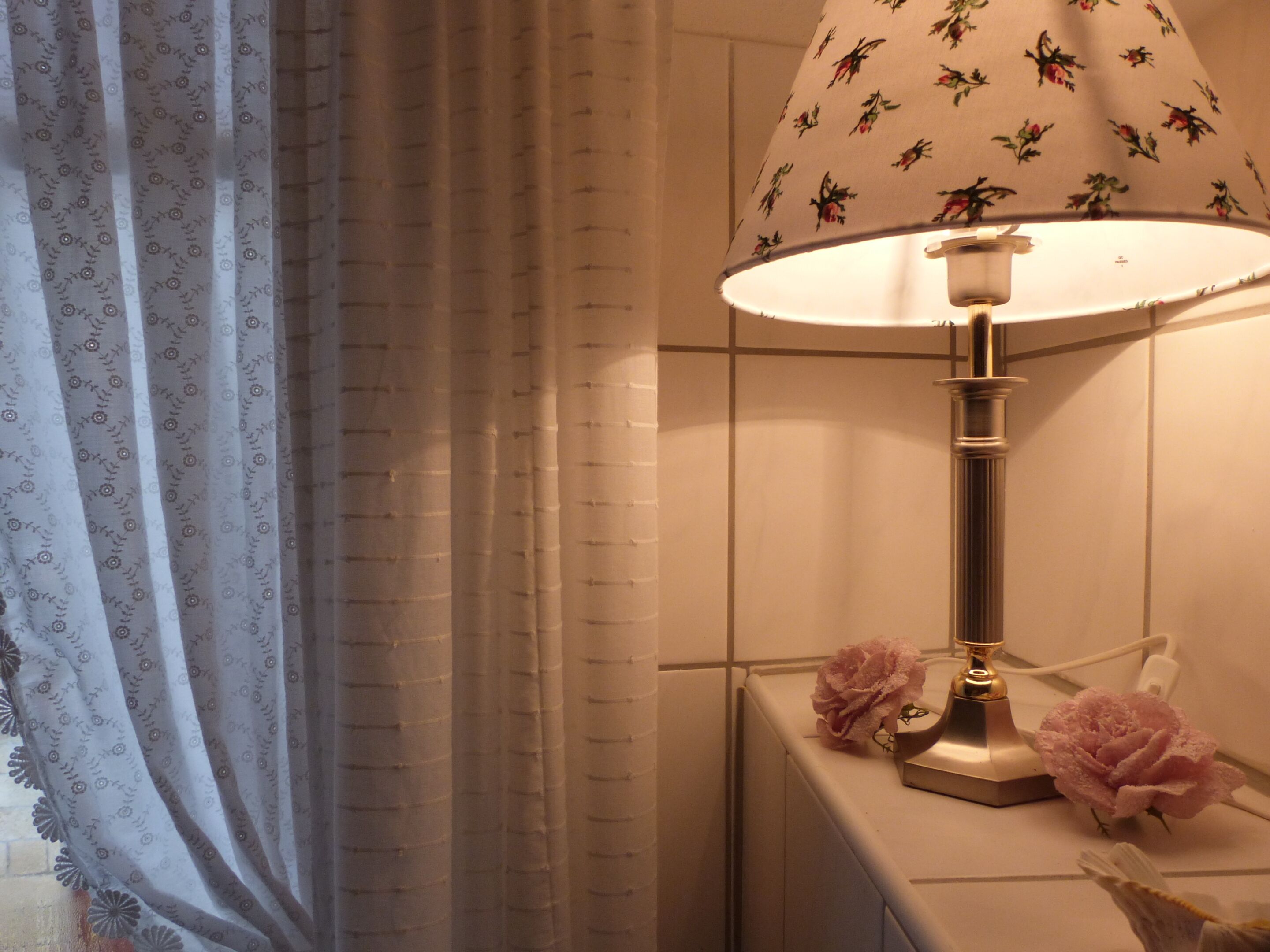 A bathroom with a white tiled wall, a floral-patterned shower curtain, a white pedestal sink with a gold faucet, and two pink flowers on the sink.